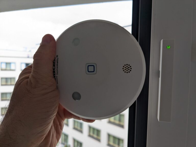 Hand hält weißes, rundes Smart-Home-Gerät mit Knopf, LEDs und Lautsprecher, rechts ein Fensterkontakt mit grüner LED, im Hintergrund ein Fenster und Mehrfamilienhaus